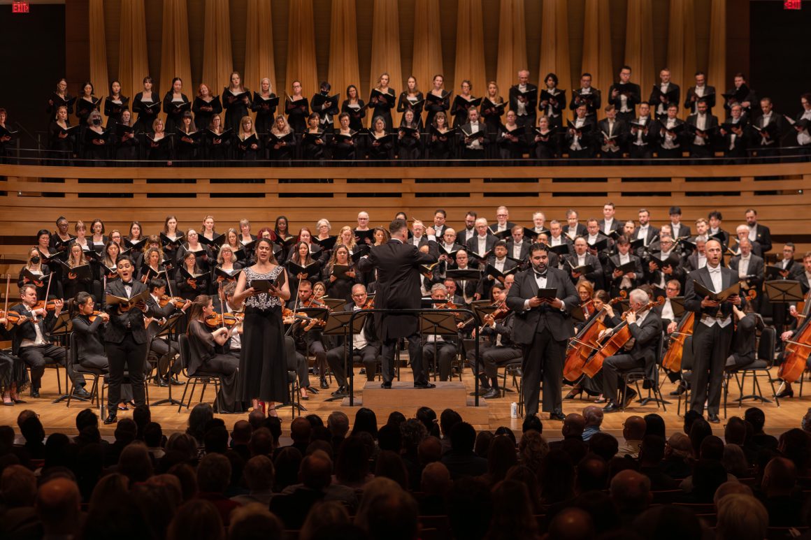Large choir and orchestra performing onstage with a conductor